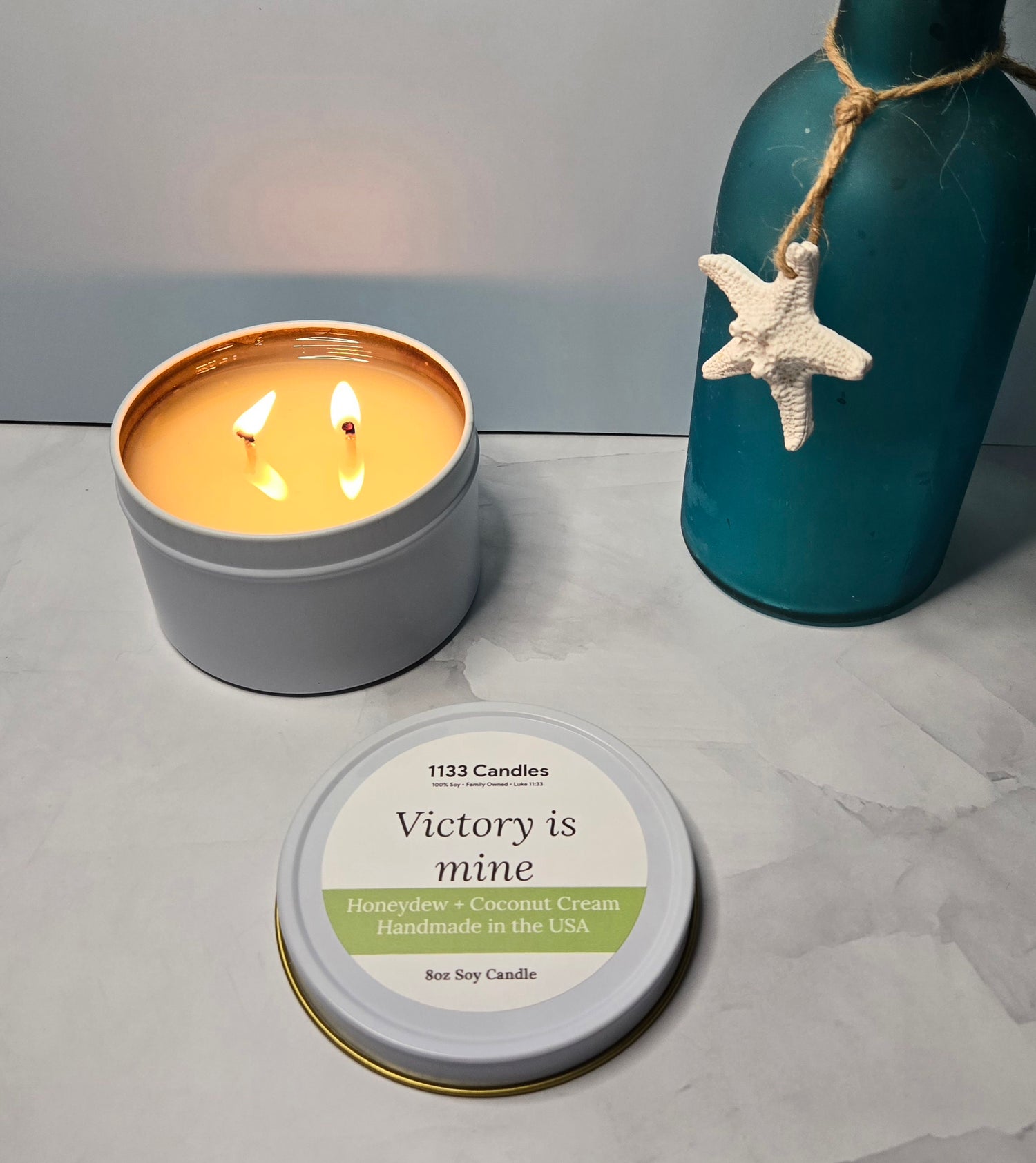 Candle in a tin with 'Victory is mine' label, surrounded by decorative items on a gray surface.